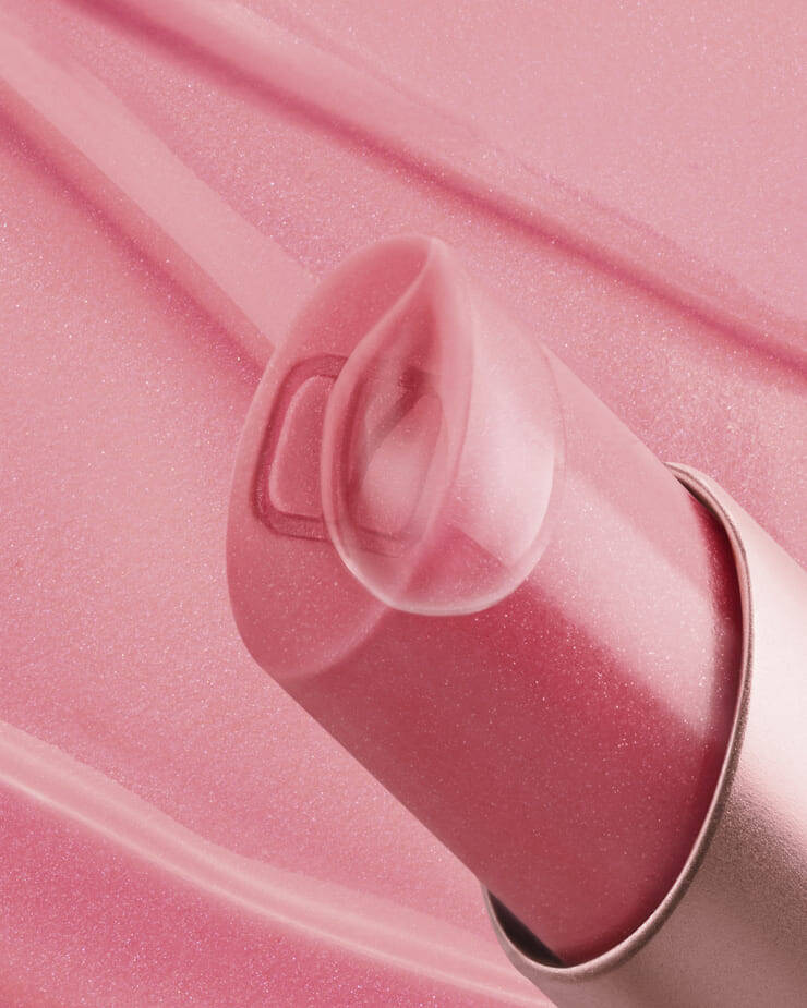 Una toma de cerca de un balsamo labial rosa, con su tapa transparente y translucida ligeramente abierta, revelando un sutil logo "V" en el interior, todo sobre un fondo rosa suave con ligeras manchas de producto.