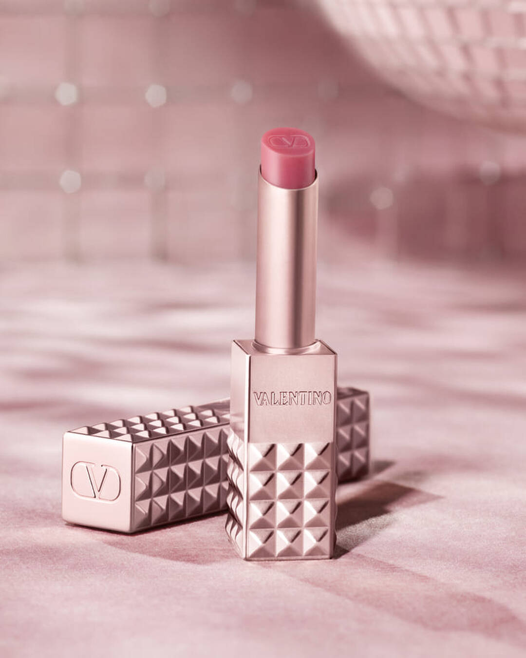 Un producto labial Valentino Spike tono oro rosa con una punta rosa se presenta en posicion vertical, con su estuche con tachuelas a juego a su lado. Ambos articulos estan colocados sobre un fondo rosa suave y brillante que evoca una sensacion de glamour.